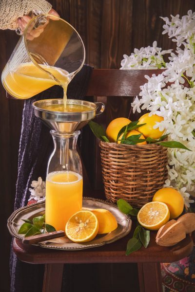 Pouring fresh orange juice from jug into glass bottle. Still life with orange juice bottle placed with sliced oranges, wooden squeezer and white flowers on rustic wooden chair.